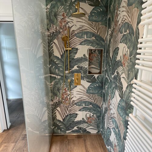 Dusche mit wunderschöner Keramik mit modernstem Aufdruck eines farbenfrohem Dschungels und goldenen Duscharmaturen. Die bodenebene Dusche wurde mit passendem Feinsteinzeug in Holzoptik gemacht um eine warme Atmosphäre zu schaffen.