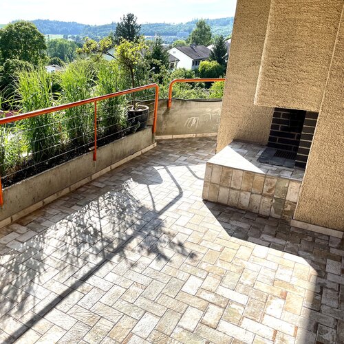 Grosser Balkon mit rustikalen kleinen Bodenplatten im Fischgrad verlegt. Die leicht grüne Farbe passt wunderbar zu den mit viel grün bepflanzen Bakonkästen.