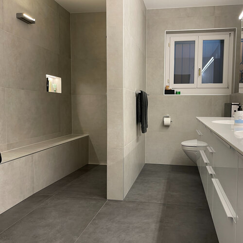 Grosszügiges Badezimmer mit Bodengleicher Dusche, einer beleuchteten Wandnische und einer Sitzbank. Unter den Fliesenbelägen sind Heiznetze eingebaut welche für warme Füsse sorgen und auch beim sitzen auf der Bank. Die modernen Grossformat Fliesen in leichter Betonoptik wurden mit viel Liebe zum Detail verlegt. 